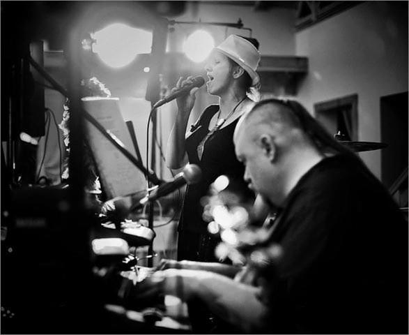 Czarno-białe zdjęcie: wokalistka z mikrofonem w kapeluszu śpiewa, pianista gra na keyboardzie, oświetlenie sceniczne w tle.