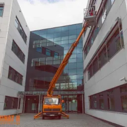 Żółty podnośnik koszowy na tle nowoczesnego budynku biurowego z przeszkloną fasadą, pracownik w koszu na wysokości okna, widoczne kamery monitoringu na elewacji.