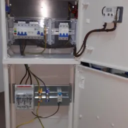 Otwarta skrzynka bezpiecznikowa z widocznymi bezpiecznikami, przewodami i licznikiem energii elektrycznej; widoczne elementy montażowe na szynie DIN.