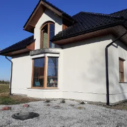 Jasny dom jednorodzinny z ciemnym dachem, widok z boku na elewację z oknami z drewnianą stolarką, na pierwszym planie szary żwir i małe rośliny.