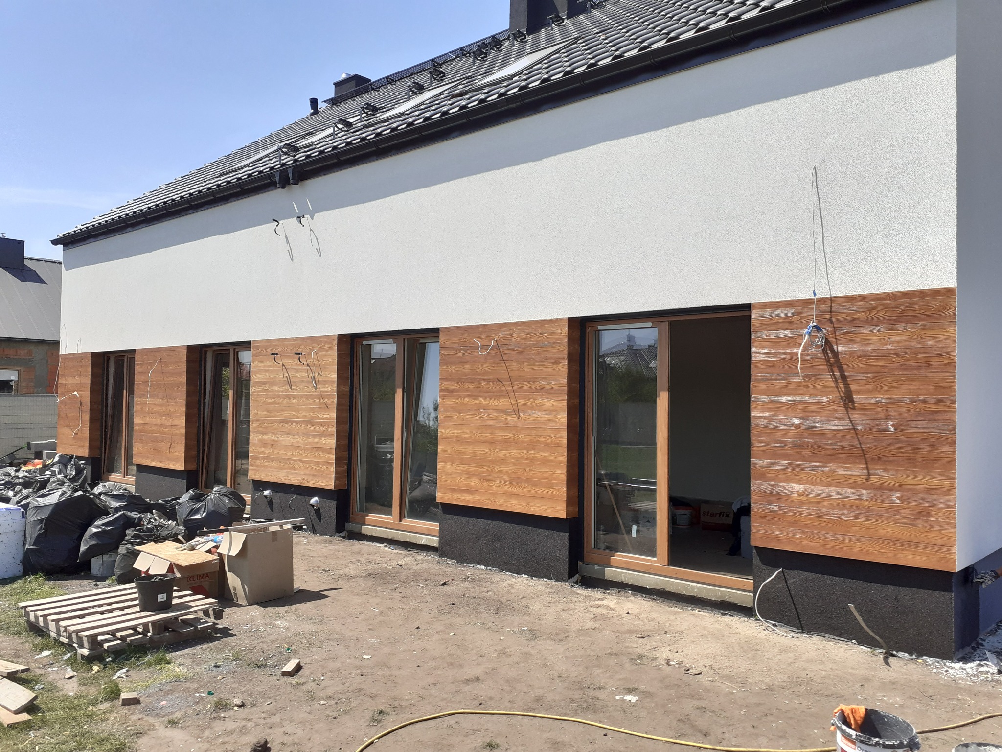 Fragment nowo wykończonej elewacji domu z drewnianą okładziną i białym tynkiem, widoczne okna, paleta z workami i kartonami, teren wokół w trakcie prac wykończeniowych.