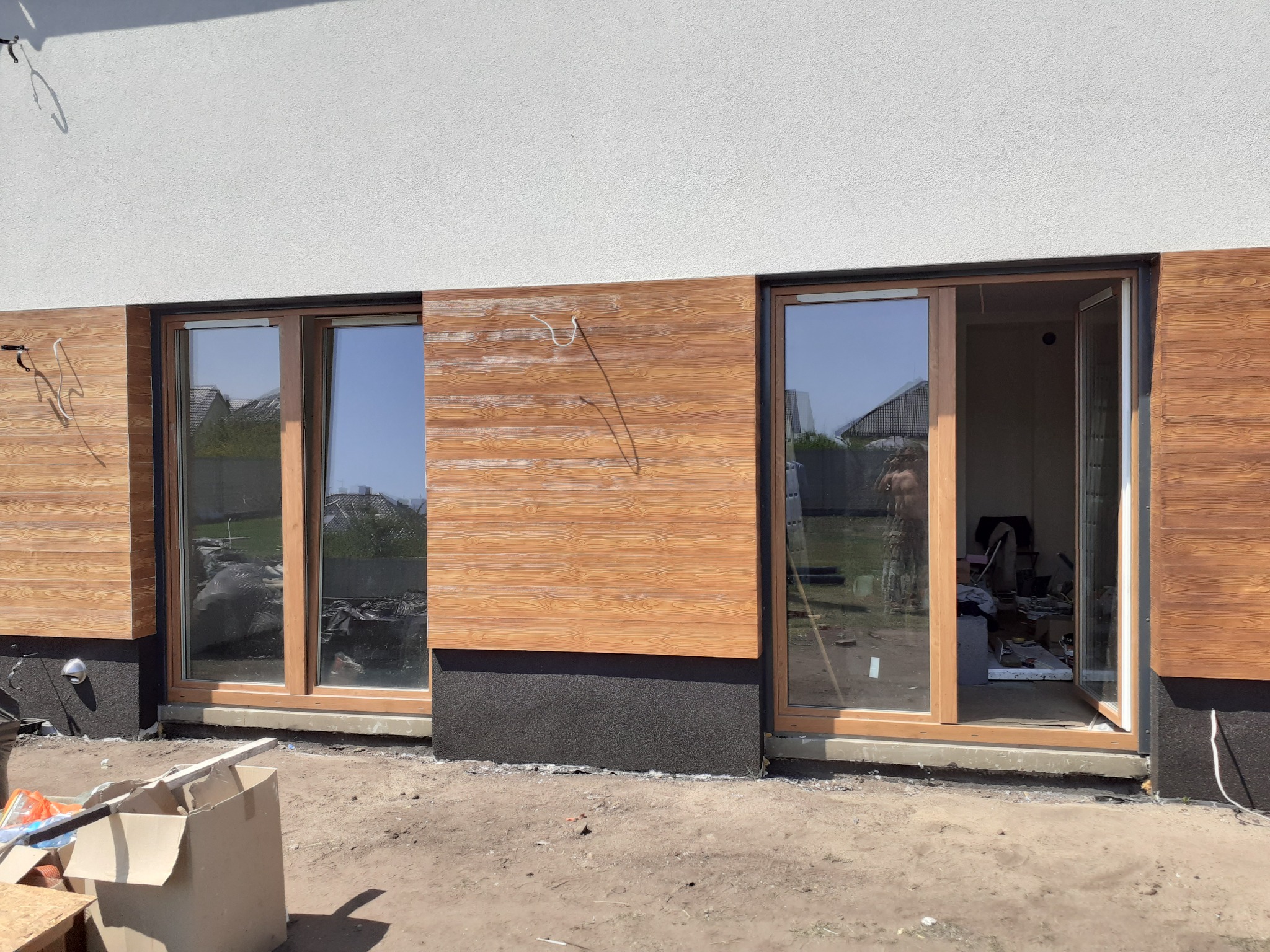 Fragment elewacji domu z drewnopodobną okładziną i dużymi oknami przesuwnymi; widoczne przygotowania do dalszych prac wykończeniowych.