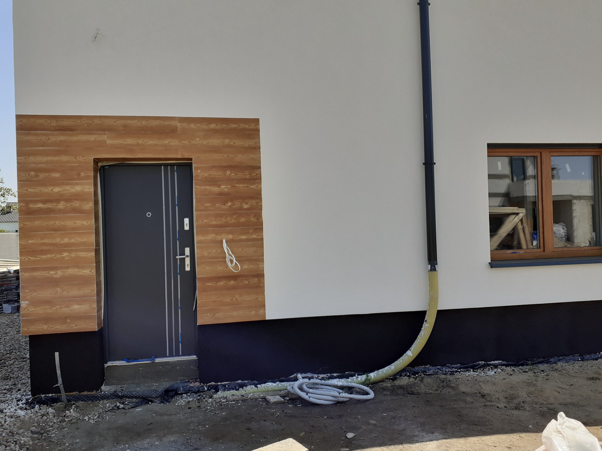 Elewacja domu w trakcie prac: widoczna ocieplona ściana z oknem, wejście z drewnopodobną okładziną, czarna izolacja fundamentów i rura do wdmuchiwania izolacji.