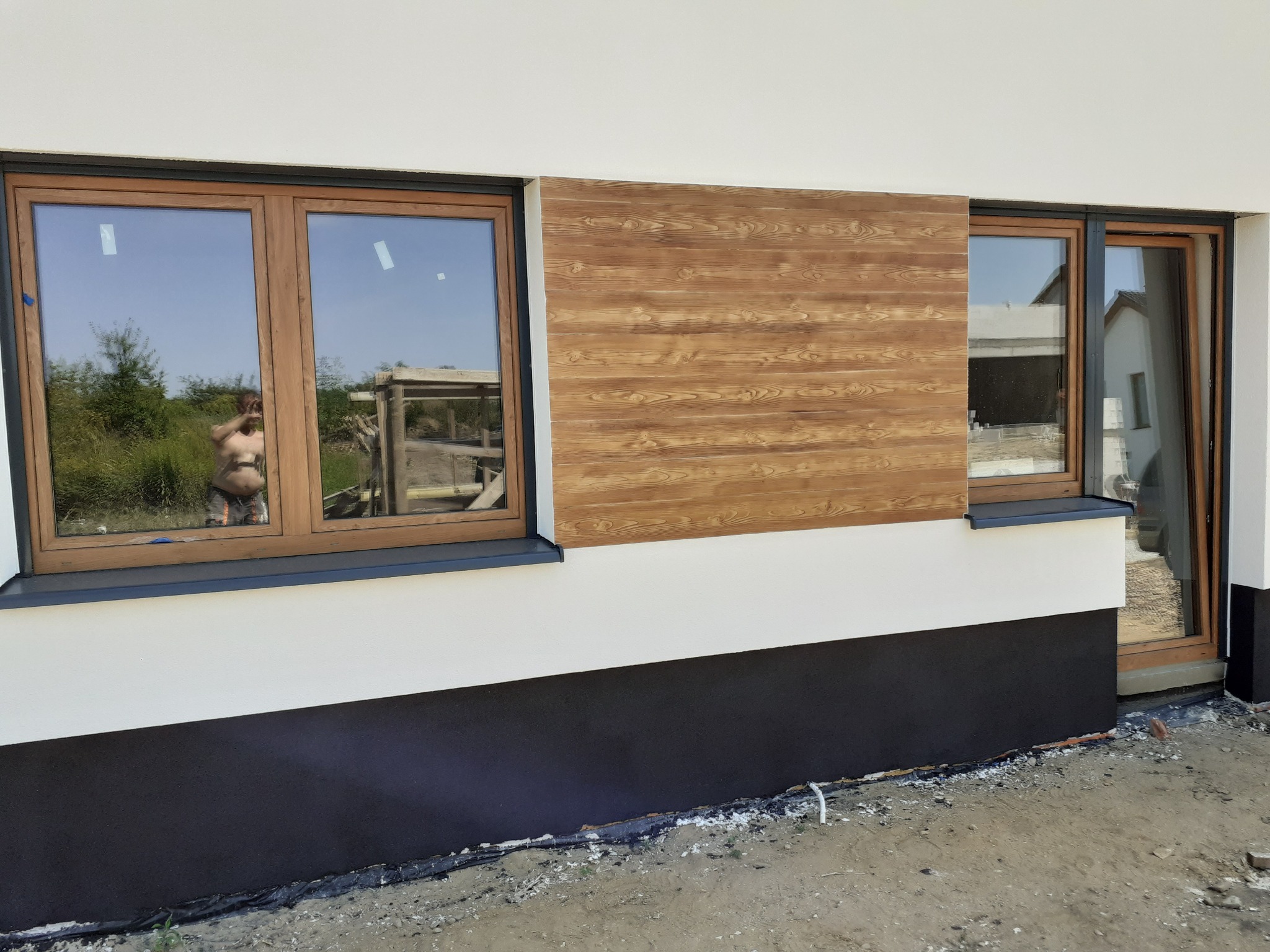 Fragment elewacji domu z drewnopodobną okładziną, widoczne dwa okna z drewnianą ramą, w jednym odbija się osoba robiąca zdjęcie, dolna część elewacji pomalowana na ciemny kolor, teren wokół domu...