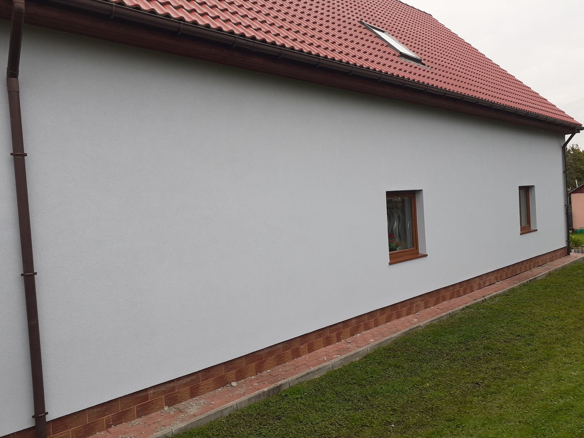 Świeżo otynkowana ściana domu jednorodzinnego z brązowymi oknami i czerwoną dachówką, widoczne fragmenty trawnika i chodnika z kostki.
