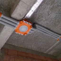 Fragment instalacji wentylacyjnej z szarymi rurami i pomarańczową skrzynką rozdzielczą, zamontowanej pod surowym stropem z widoczną izolacją i fragmentem murowanej ściany.