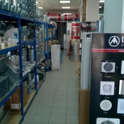 Długi korytarz magazynu z półkami pełnymi metalowych rur wentylacyjnych i materiałów izolacyjnych Rockwool. Na końcu widoczny szyld DOSF z wentylatorami.