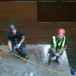 Dwóch pracowników wysokościowych, jeden w żółtej kamizelce i czerwonym kasku, drugi w czarnym ubraniu i szarym kasku, przygotowuje się do pracy na elewacji budynku o brązowej, panelowej strukturze,...