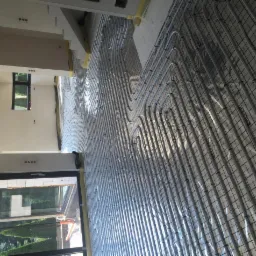 Ułożone rury ogrzewania podłogowego na siatce w jasnym pomieszczeniu z oknem. Widoczne elementy instalacji i przygotowanie do zalania posadzką.