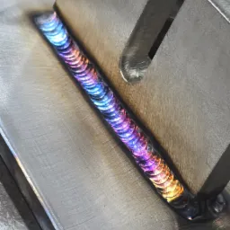 Szlif spawalniczy na metalowej płycie z widocznymi kolorami tęczy, przymocowany metalowymi klockami.