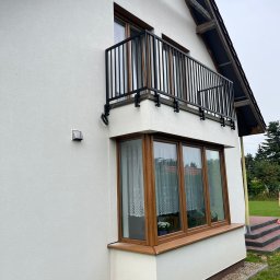 Usługi Spawalnicze Jarosław Kwiecień - Biały dom z czarną balustradą balkonową o prostym wzorze, drewnianymi oknami i wnęką okienną z kwiatami za firanką.