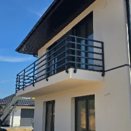 Nowoczesny dom z balkonem zabezpieczonym czarną, stalową balustradą o prostym, geometrycznym wzorze, z widoczną koparką Volvo w tle, sugerującą prace budowlane lub wykończeniowe.