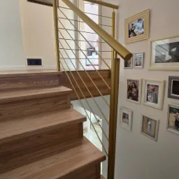 Złota balustrada ze stali nierdzewnej z poziomymi prętami, zamontowana przy drewnianych schodach w jasnym wnętrzu z galerią zdjęć na ścianie.