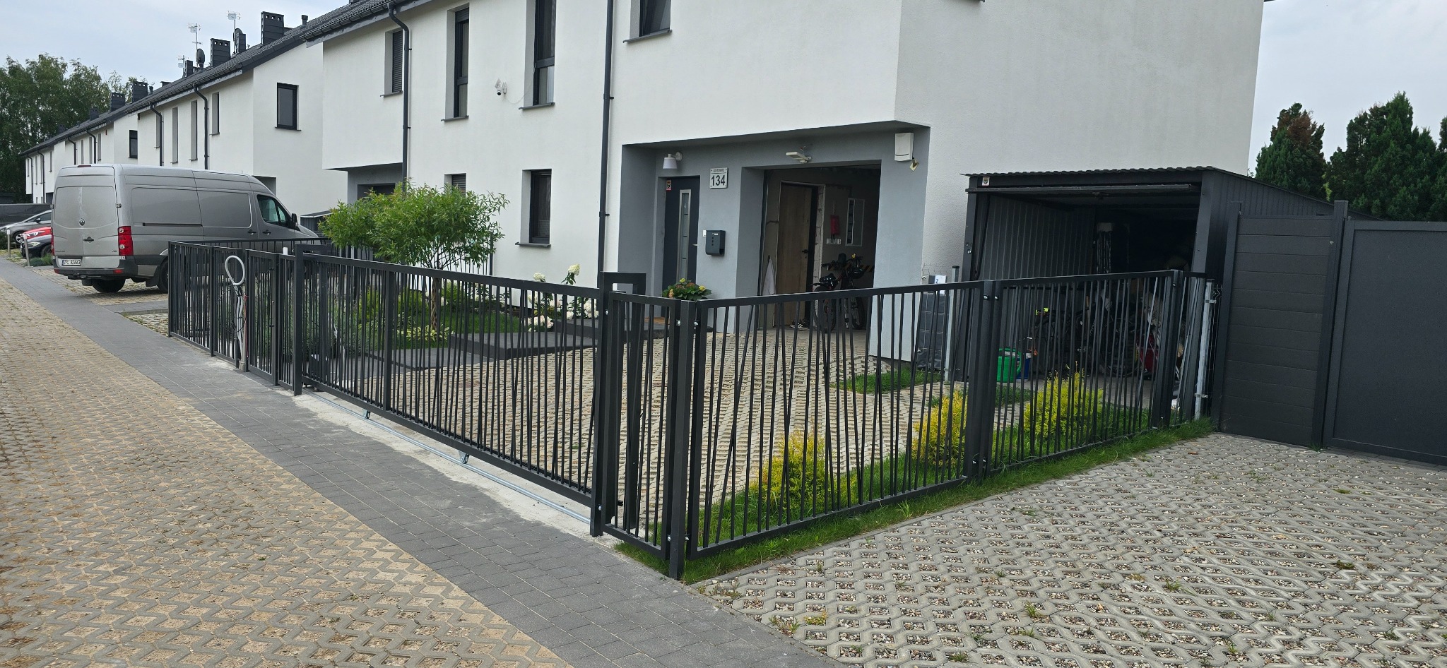 Nowoczesne, czarne ogrodzenie metalowe z pionowych prętów, oddzielające posesję od chodnika wyłożonego ażurową kostką; w tle białe domy szeregowe i zaparkowany van.