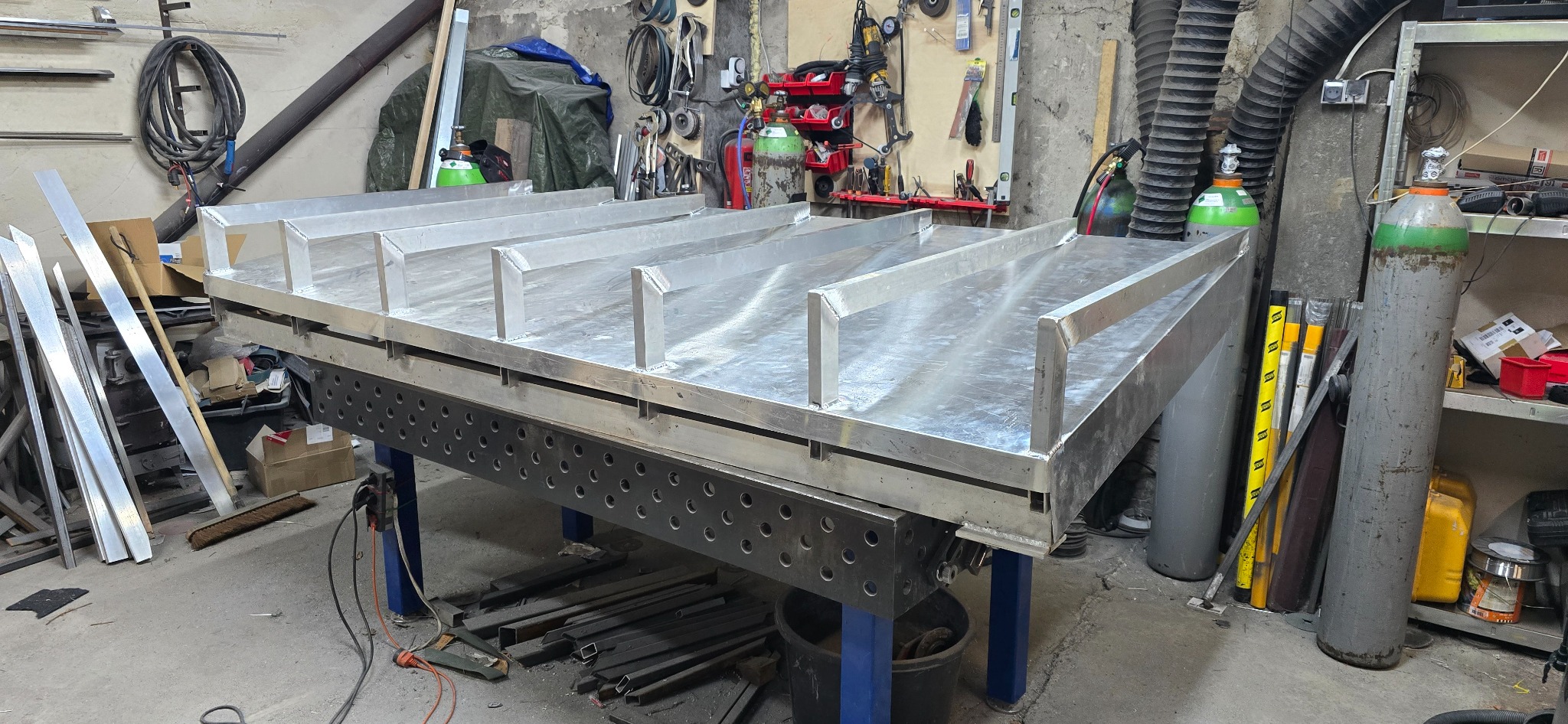 Konstrukcja spawana z profili aluminiowych na stole spawalniczym z otworami, widoczne butle z gazem i narzędzia w tle warsztatu.