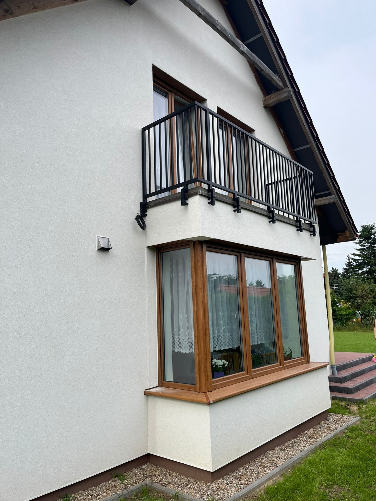 Biały dom z czarną balustradą balkonową o prostym wzorze, drewnianymi oknami i wnęką okienną z kwiatami za firanką.