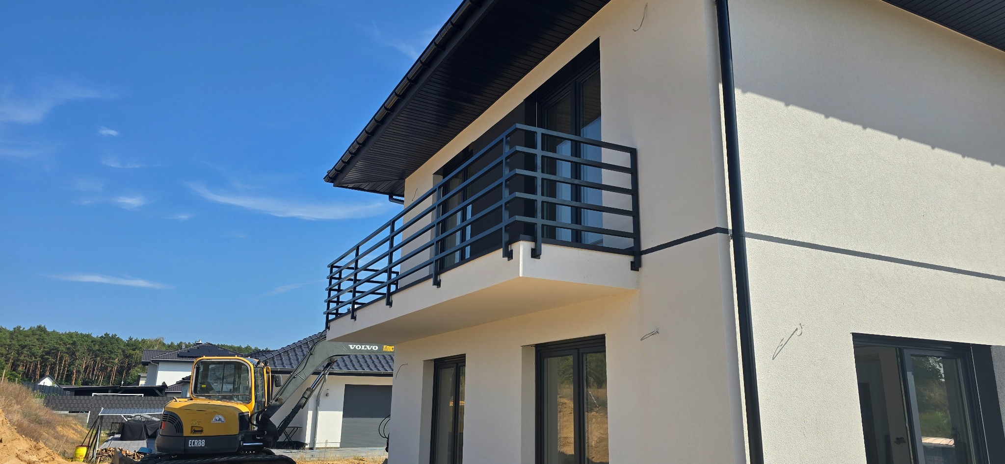 Nowoczesny dom z balkonem zabezpieczonym czarną, stalową balustradą o prostym, geometrycznym wzorze, z widoczną koparką Volvo w tle, sugerującą prace budowlane lub wykończeniowe.