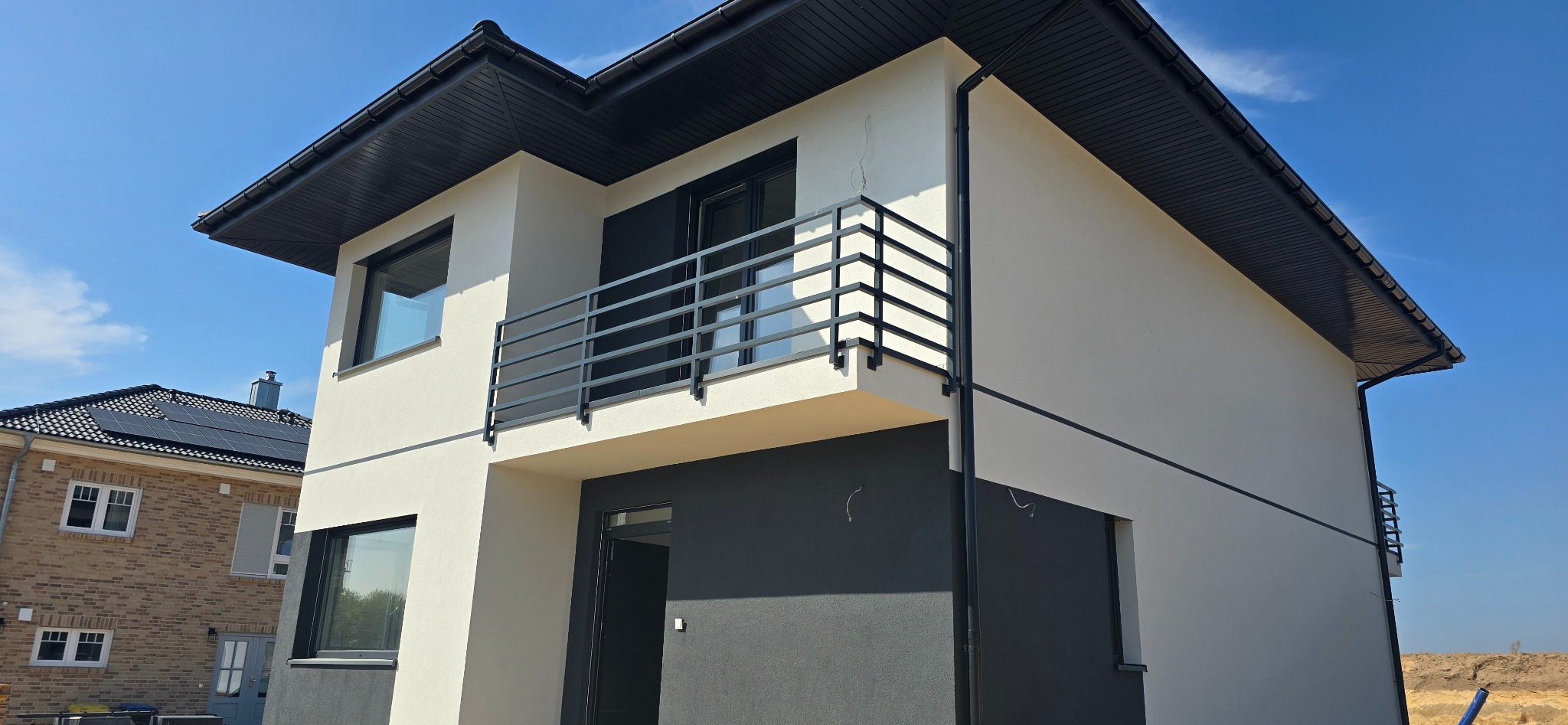 Nowoczesny dom z balkonem zabezpieczonym prostą, czarną balustradą poziomą. Widoczne okna z czarnymi ramami, fragment dachu i elewacja w odcieniach bieli i szarości. W tle błękitne niebo.