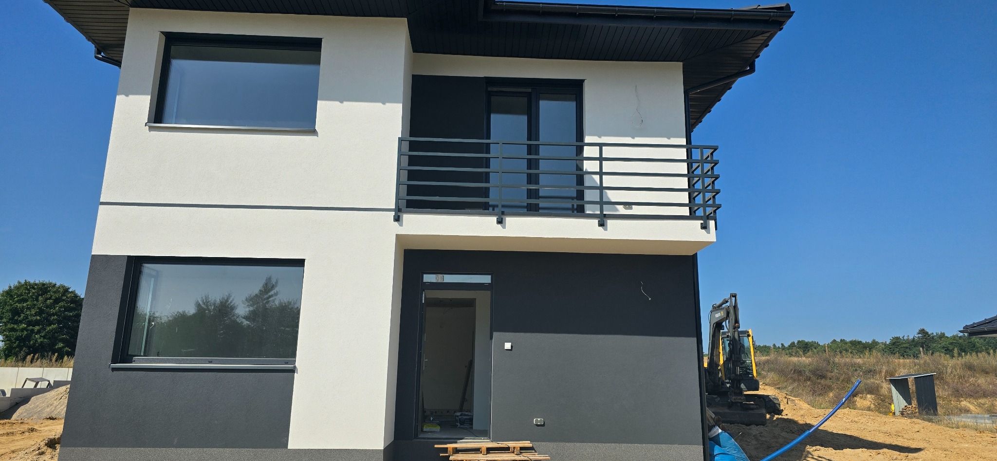 Dwukondygnacyjny dom z balkonem zabezpieczonym czarną, prostą balustradą. Widoczny fragment elewacji w kolorze białym i szarym, okna z czarnymi ramami i drzwi balkonowe. Na pierwszym planie paleta...