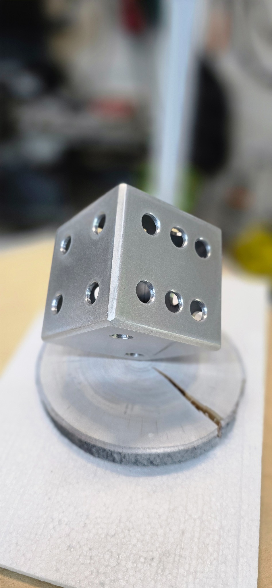 Metalowy sześcian z otworami, przypominający kostkę do gry, umieszczony na okrągłej, metalowej podstawie z widocznym pęknięciem.