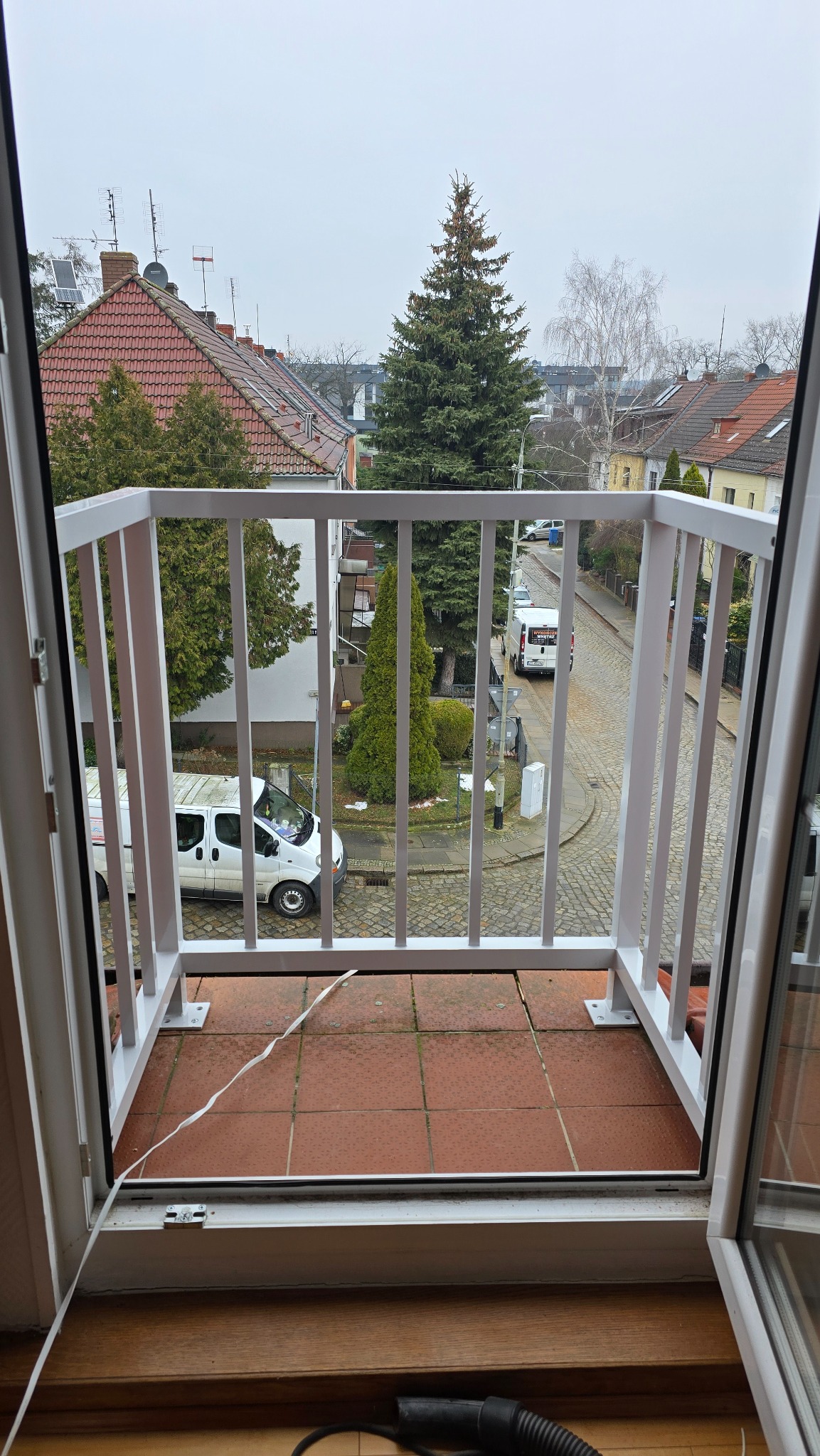 Widok z balkonu z białą, metalową balustradą na ulicę z brukowaną nawierzchnią i zabudową mieszkalną w tle, z widocznym fragmentem wnętrza z brązową podłogą i odkurzaczem.