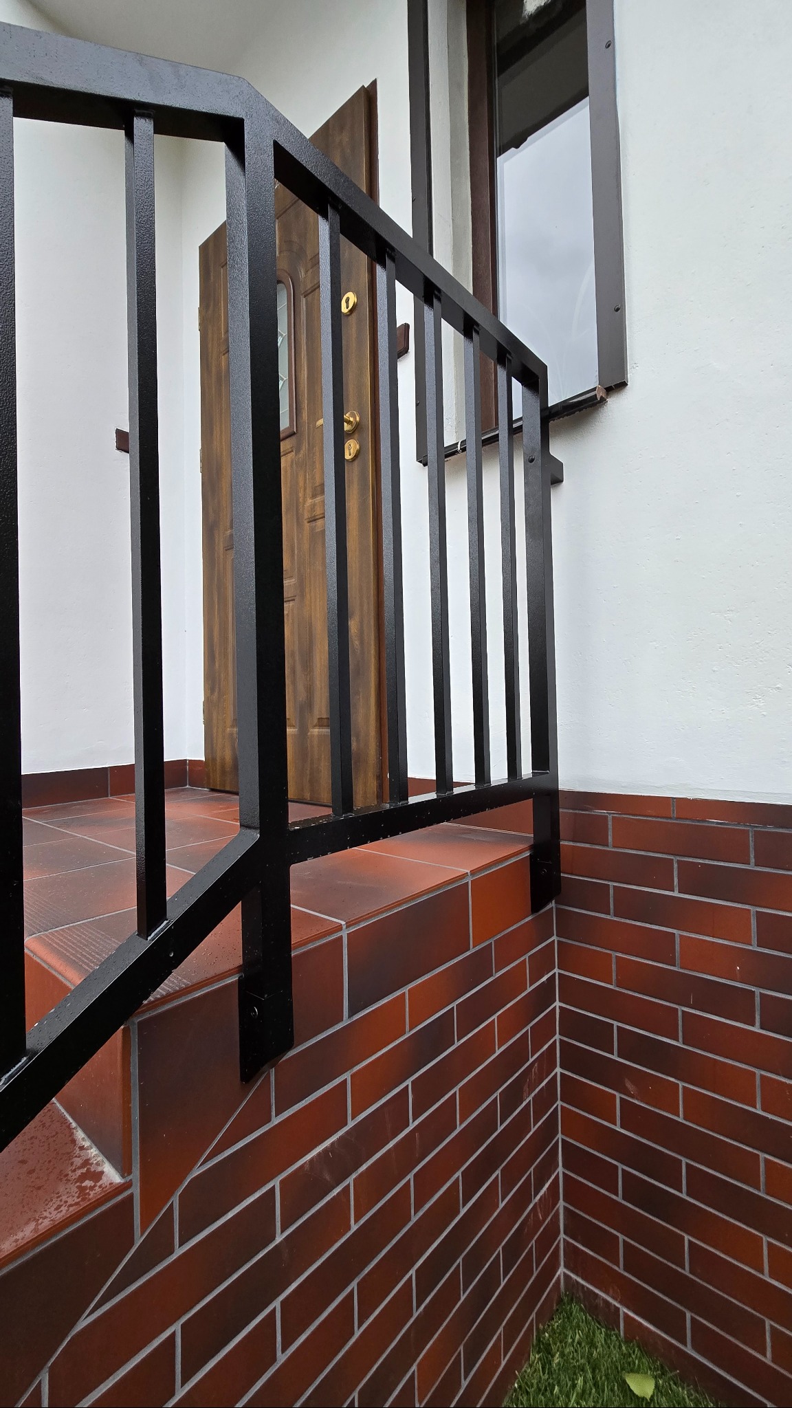 Czarna, metalowa balustrada schodowa z pionowymi szczeblami na tle białej ściany i drewnianych drzwi wejściowych. Widok z dołu, podkreślający solidność konstrukcji.