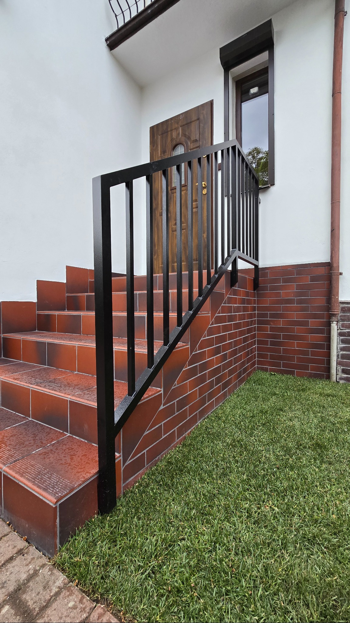 Czarna, aluminiowa balustrada schodowa z maskownicami śrub, malowana proszkowo. Schody wyłożone czerwoną terakotą, obok trawnik i ceglana elewacja.