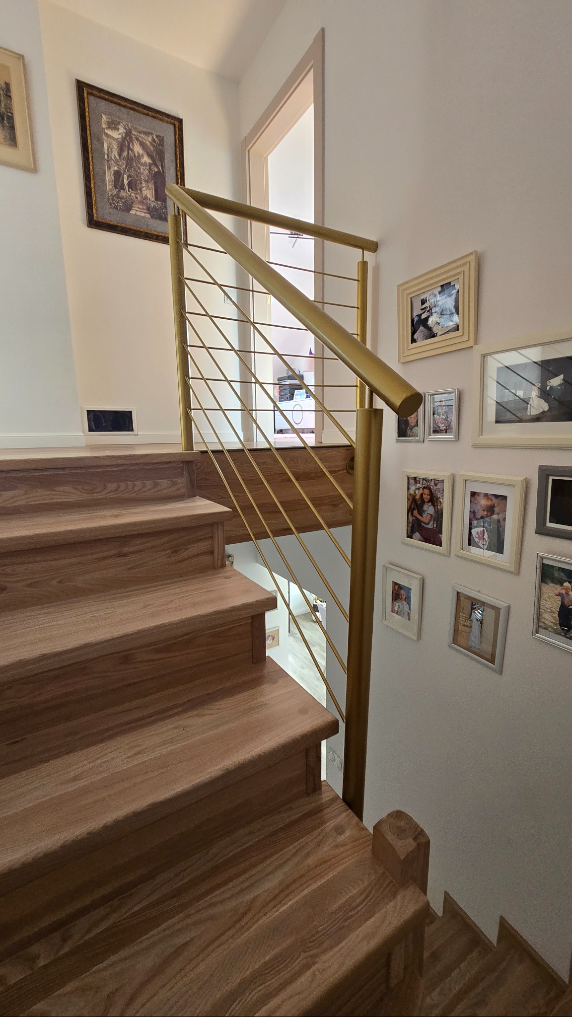Złota balustrada ze stali nierdzewnej z poziomymi prętami, zamontowana przy drewnianych schodach w jasnym wnętrzu z galerią zdjęć na ścianie.