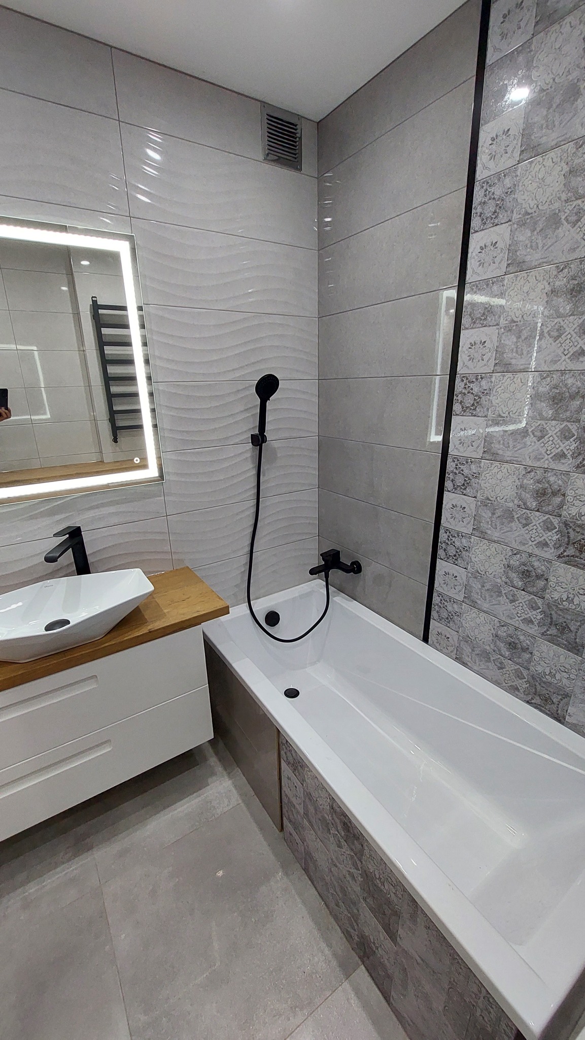 Nowoczesna łazienka z białą, prostokątną umywalką nablatową, drewnianym blatem, czarną armaturą, wanną obudowaną płytkami imitującymi kamień oraz płytkami ściennymi w odcieniach szarości z różnymi...