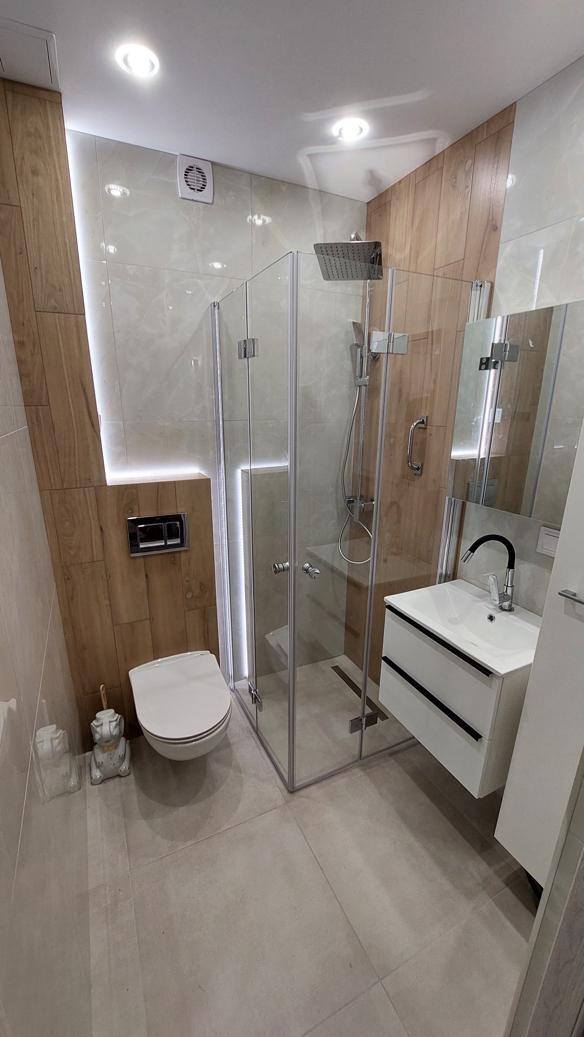 Nowoczesna łazienka z kabiną prysznicową typu walk-in, wiszącą toaletą, umywalką z czarnymi akcentami i drewnopodobnymi płytkami ściennymi, oświetlona listwą LED.