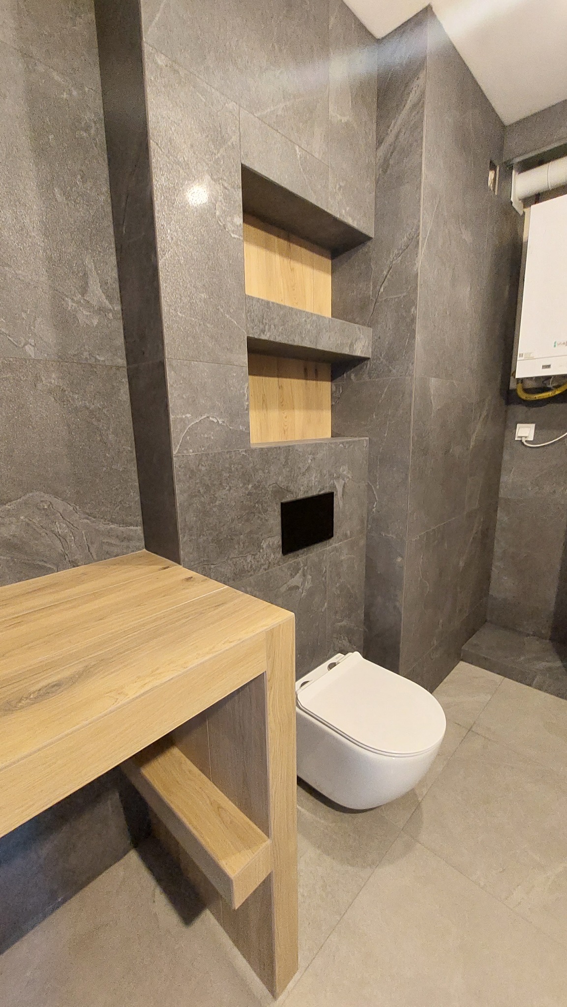 Szare płytki imitujące kamień na ścianie i podłodze, wisząca toaleta, drewniany blat z półką, wnęki z drewnianym wykończeniem, czarny panel sterujący spłuczką.