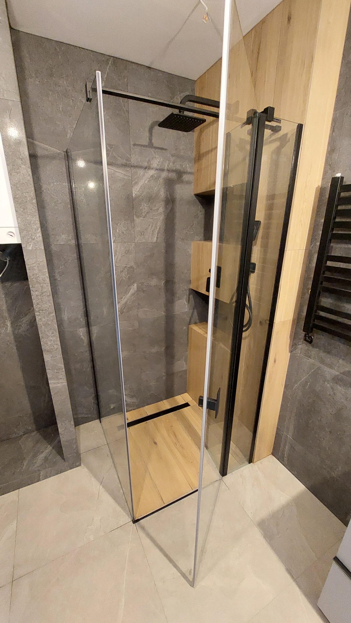 Nowoczesna kabina prysznicowa typu walk-in z czarnymi profilami, drewnianym wykończeniem ściany i odpływem liniowym, szare płytki na ścianach, beżowe płytki podłogowe.