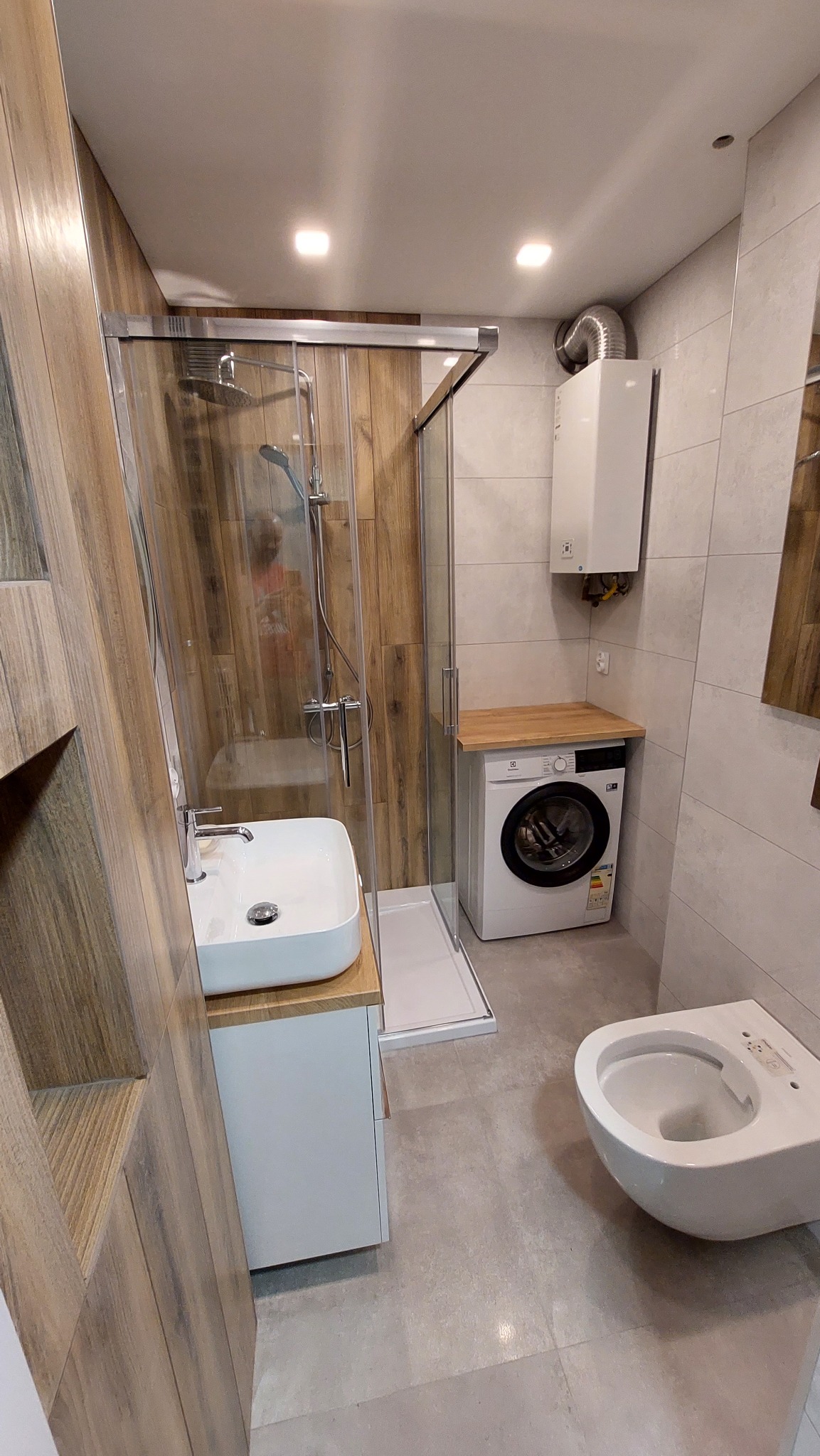 Nowoczesna łazienka z kabiną prysznicową typu walk-in, umywalką nablatową na białej szafce, pralką pod blatem z bojlera oraz podwieszaną miską WC, utrzymana w szaro-beżowej kolorystyce z akcentami...