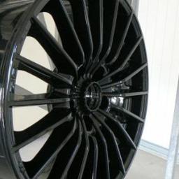 Dom-Wid - Błyszcząca, czarna felga aluminiowa z wieloma ramionami, po malowaniu proszkowym, zawieszona na metalowym stelażu w hali produkcyjnej.