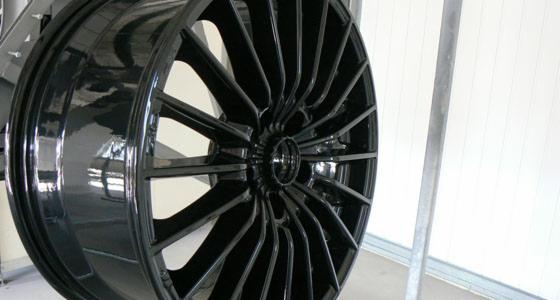 Błyszcząca, czarna felga aluminiowa z wieloma ramionami, po malowaniu proszkowym, zawieszona na metalowym stelażu w hali produkcyjnej.