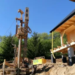 Wiertnica na gąsienicach podczas wiercenia studni głębinowej obok domu w trakcie budowy, widoczny żółty agregat prądotwórczy Beretta T44/GT.