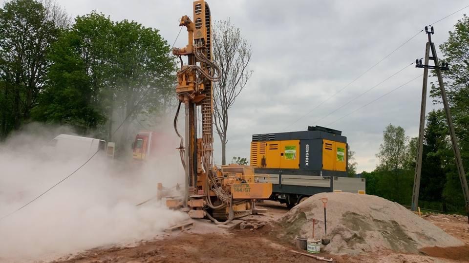 Żółta wiertnica T44/GT w akcji, obok agregat XRH5, unoszący się pył, drzewa w tle. Studnia głębinowa w trakcie wiercenia.