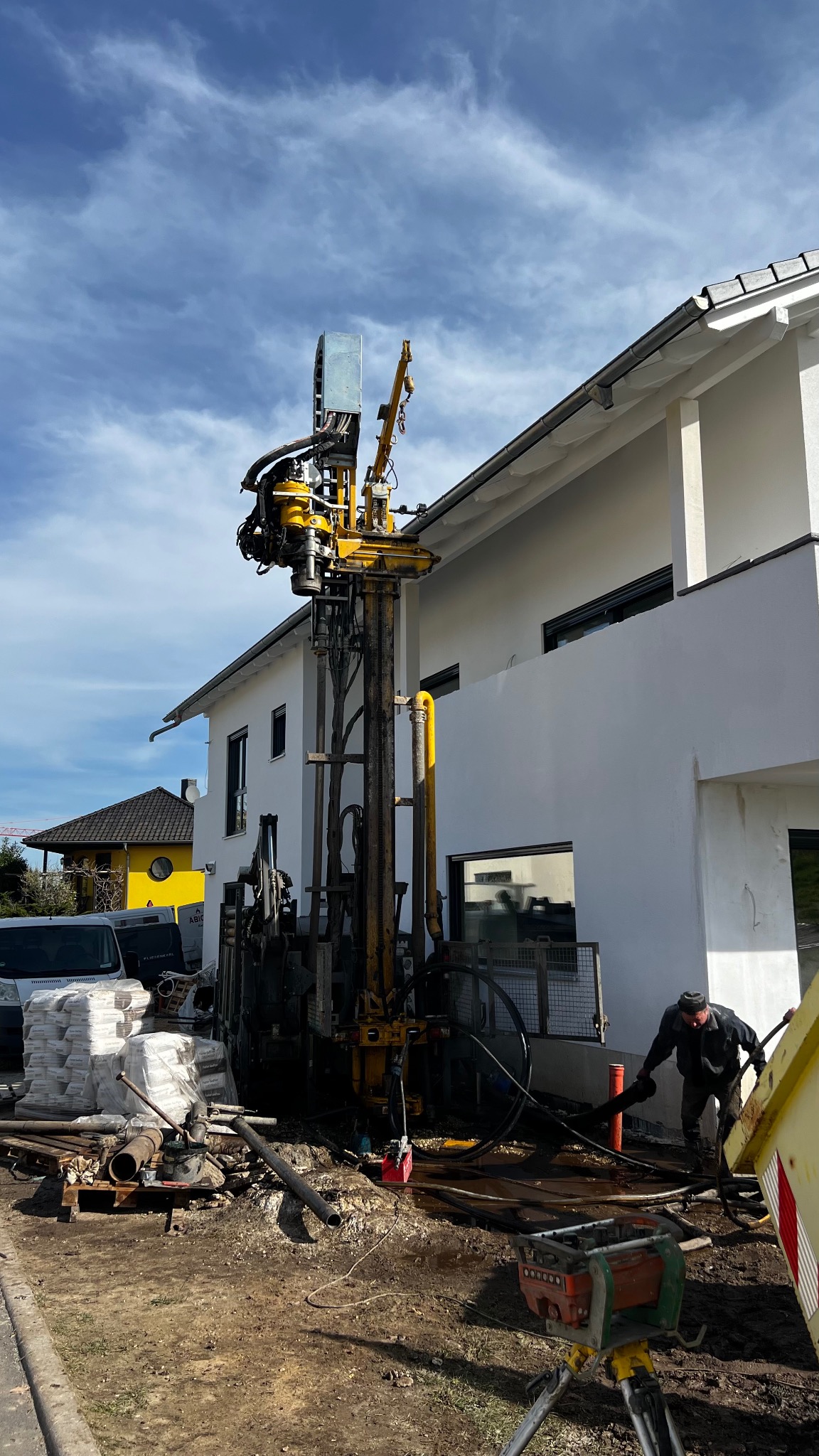 Wiertnica do studni głębinowych w bliskiej odległości od białego domu z szarym dachem. Operator w trakcie pracy. Worki z cementem na palecie obok.