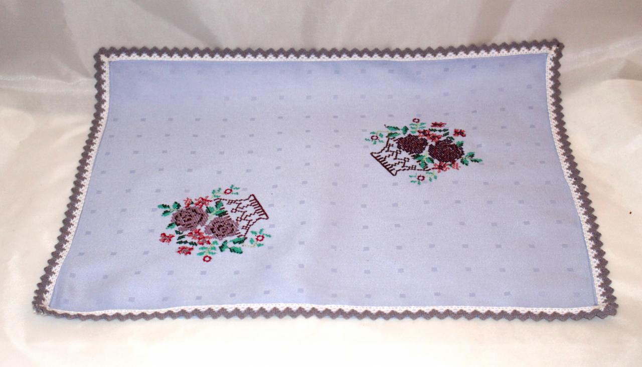 Ręcznie haftowana serweta w pastelowym odcieniu błękitu, zdobiona motywem kwiatów w koszyczkach i wykończona szarą koronką.