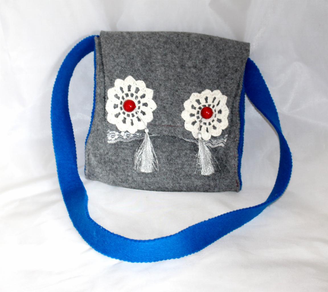 Ręcznie szyta, szara torebka z filcu, ozdobiona koronkowymi kwiatami z czerwonymi koralikami i srebrnymi frędzlami, na niebieskim pasku, prezentowana na białym tle.