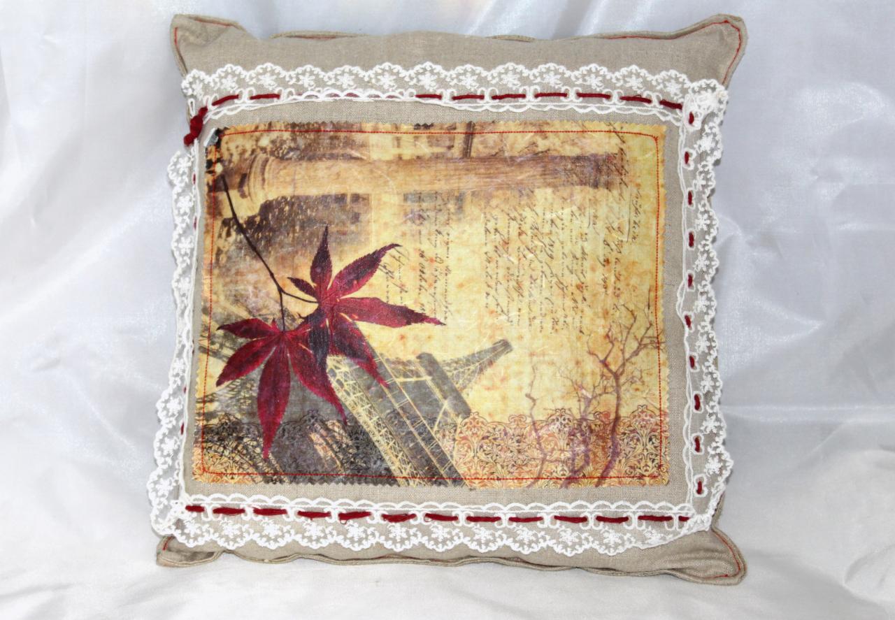 Dekoracyjna, beżowa poduszka z motywem wieży Eiffla i czerwonych liści klonu, obszyta białą koronką i czerwoną nitką. Idealna jako element wystroju wnętrza.