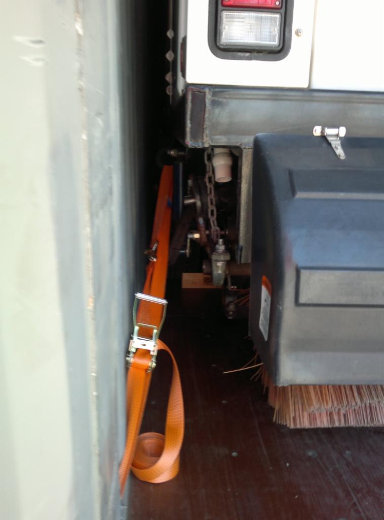 Wnętrze skrzyni ładunkowej ze szczotką zamiatarki przymocowanej pomarańczowym pasem transportowym do ściany. Widoczny mechanizm łańcuchowy i tylne światło pojazdu.