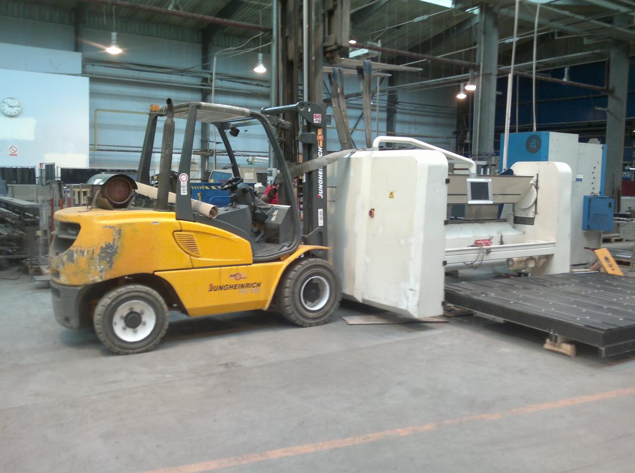 Żółty wózek widłowy Jungheinrich transportuje element maszyny CNC w hali produkcyjnej. Widoczne oświetlenie przemysłowe i elementy wyposażenia.