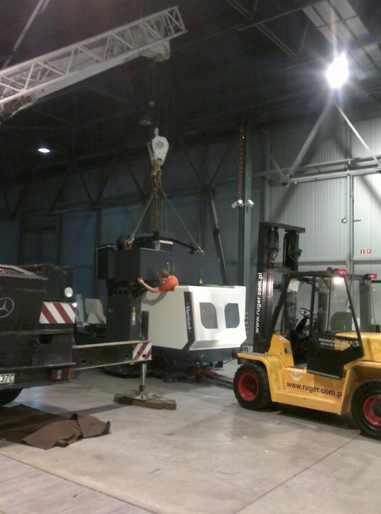 Montaż maszyny Hartford w hali produkcyjnej z użyciem dźwigu i wózka widłowego. Pracownik w pomarańczowej koszulce nadzoruje proces.