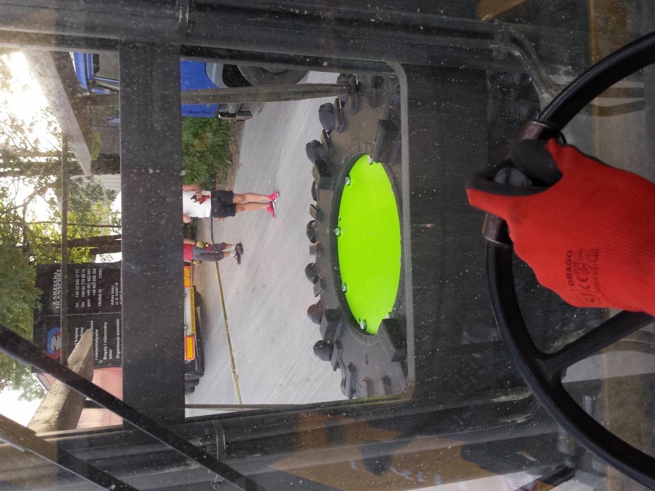 Wnętrze kabiny maszyny rolniczej z widokiem na zielone koło i osoby na zewnątrz. Ręka w czerwonej rękawicy na kierownicy, brudna szyba.