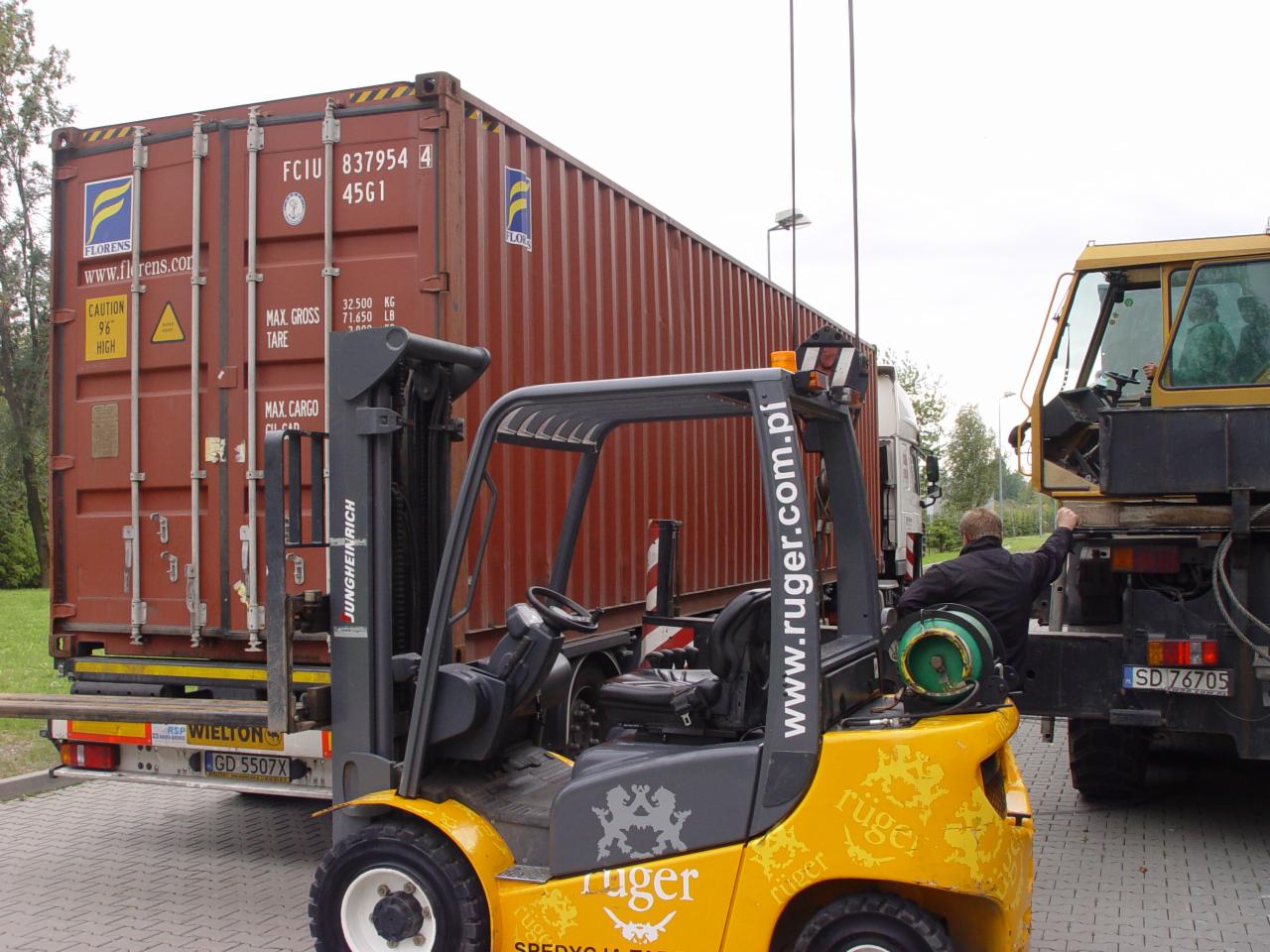 Żółty wózek widłowy Jungheinrich obok kontenera na naczepie, z widocznym fragmentem dźwigu i operatorem. Ruger.com.pl na boku wózka.