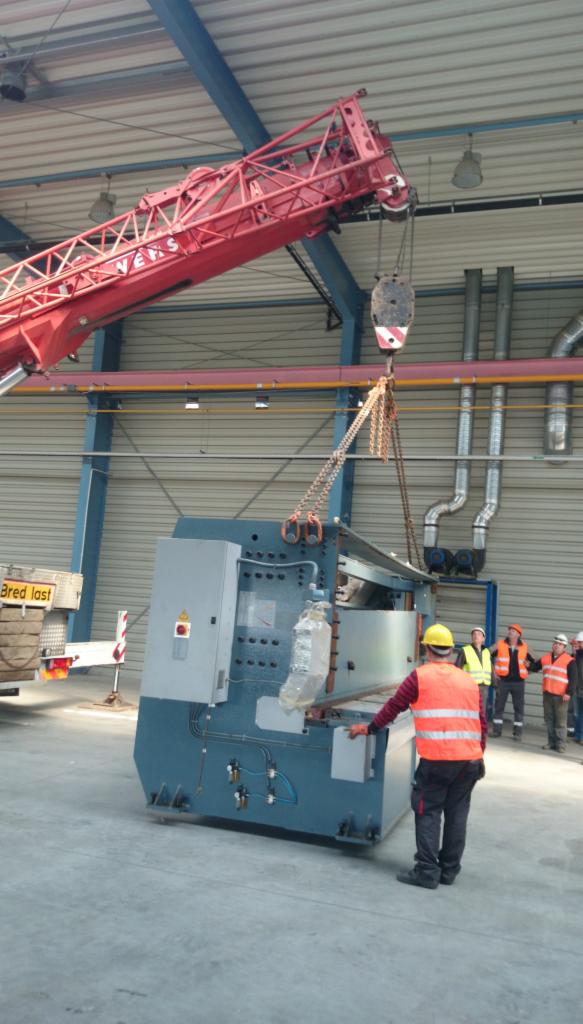 Transport maszyny przemysłowej w hali za pomocą czerwonego dźwigu. Grupa osób obserwuje proces. Widoczne elementy instalacji przemysłowej.