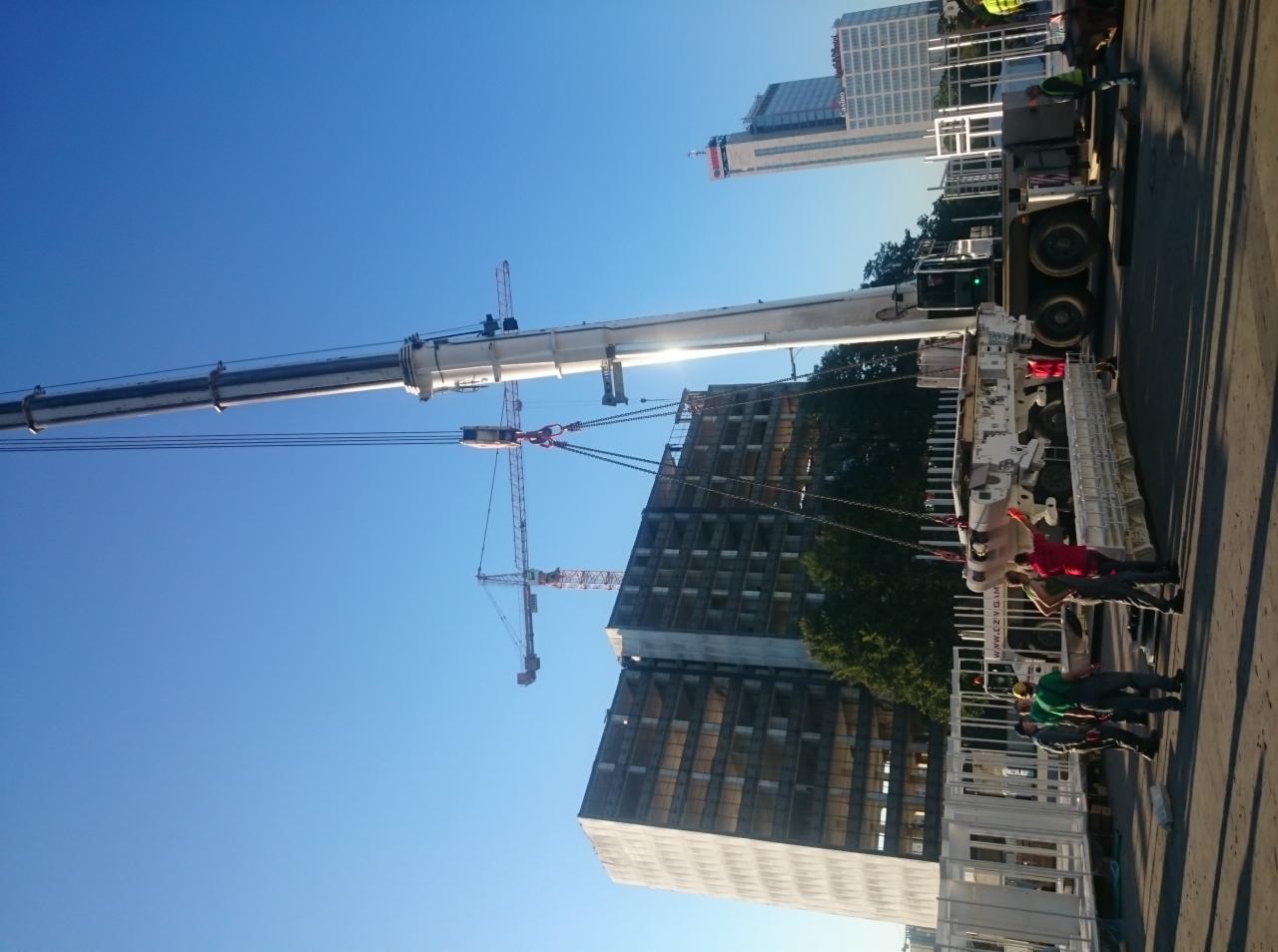 Żuraw wieżowy w akcji na budowie, widoczne budynki w tle i pracownicy na ziemi. Kadr uchwycony z niskiej perspektywy, błękitne niebo w tle.