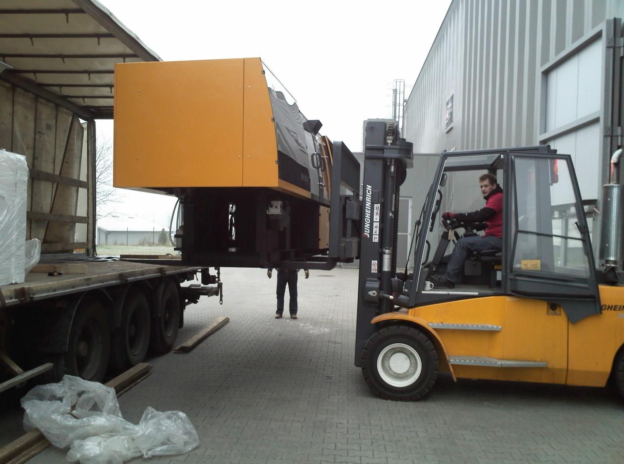 Żółty wózek widłowy Jungheinrich z operatorem transportuje dużą, żółto-czarną maszynę z naczepy ciężarówki na placu; widoczna postać w tle.