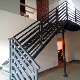 Nowoczesne schody wewnętrzne z metalową konstrukcją i balustradą z poziomych profili, prowadzące na antresolę w surowym wnętrzu z ceglanym kominem.